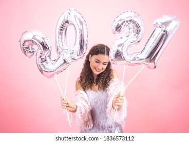 Cute young girl with a smile in a festive outfit on a pink studio background holding silver balloons from 2021 numbers