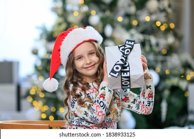 fröhliches Mädchen in Santa Red Hat mit Geschenkbox mit schwarzem Band Hintergrund Weihnachtsbaum. Weihnachtsgeschenke Konzept. Feier. Kind mit lockigem Haar und blauen Augen im Pullover. Einkaufen vor dem Urlaub
