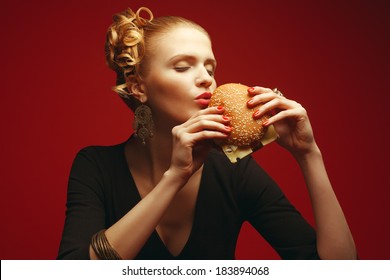 不健康な食事。ジャンクフードのコンセプト。罪深い楽しみ。赤い背景の上でチーズバーガーを持って食べるおしゃれなヒップスターの女の子。完璧な髪、肌、メイク、マニキュア。コピースペース。スタジオ撮影