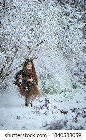 Fairy tale girl. Portrait a little girl in a deer dress with a painted face in the winter forest. Big brown antler. Fantasy girl with christmas wreath. Snowy park.