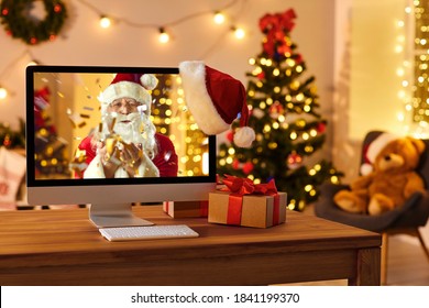Computer on table in cozy room with hanging red hat and with Santa Claus on screen blowing golden confetti, sending love, wishing Merry Christmas and Happy New Year online and making miracle come true