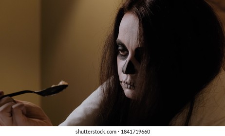Una niña con la apariencia de un lindo zombi se sienta en la cama, envuelta en una manta blanca, vacilante se lleva una cucharada de mermelada de cerebro a la boca, la prueba con la lengua y disfruta. Víspera de Halloween. De cerca.