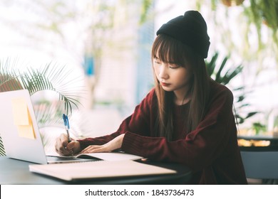 若い大人のアジアの大学生の女性は、赤いセーターとウールの帽子を着用します。ラップトップを使用して勉強し、カフェでオンラインで作業します。冬の日に家の外で大学の人々 の都市生活。