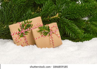 Two handmade plastic-free Christmas presents under fir tree. Gifts wrapped in craft paper and rope bow stand in snow with pine tree branches as background. Winter holiday celebration, giving presents