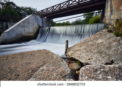 Verlassener kleiner Betonwasserfall mit einer Holzbrücke darüber und großen Betonblöcken (Felsen) vorne.