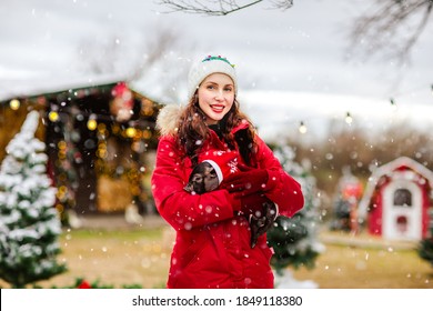 Junge attraktive Frau im roten Winteroverall und im weißen Hut, der mit dem kleinen schwarzen Schwein gegen Weihnachtshintergrund aufwirft.