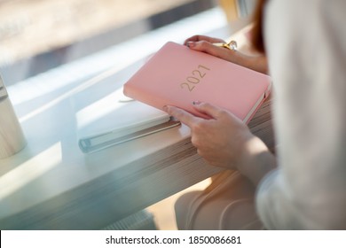 Female hands holding pink or coral coloured leather diary 2021 and pen while sitting near a window Concept of planning personal future goals and ideas for new year 2021. Copy space.