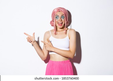 ピンクのかつらとハロウィーンの化粧をした熱狂的なパーティー ガールの画像、何か幻想的なものを見て、指を左に向け、白い背景の上に立っています。