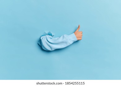 Menselijke hand die door papieren muur breekt en duim toont als teken van goedkeuring of overeenkomst geïsoleerd over blauwe studioachtergrond. Plaats voor tekstinformatie. Acceptatie oke en leuk concept