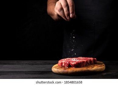 シェフは、木製のテーブルの黒い背景にまな板の上の新鮮なステーキに塩を振りかけます。水平方向の画像。ロゴ テキスト ラベル用の空き容量あり