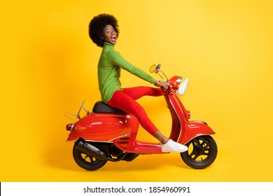 Photo portrait of excited african american woman driving a red scooter with legs spread wearing casual outfit isolated on vivid yellow colored background