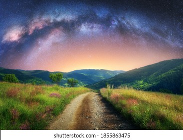 夏の山の未舗装の道路にアーチ型の天の川。星空、天の川のアーチ、山里の小道、丘、緑の草、紫の花が美しい夜景。宇宙と銀河
