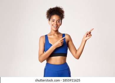 Concepto de entrenamiento y fitness. Atractiva atleta afroamericana en traje deportivo azul, señalando con el dedo a la derecha y sonriendo, mostrando publicidad, fondo blanco