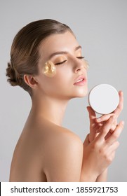 彼女の顔に花がクリームの瓶を保持するように形成された、薄いメイクと金の目パッチ マスクを持つ美しい若い女性。自然な化粧をした美容モデルのポートレートは、肌を気遣う。