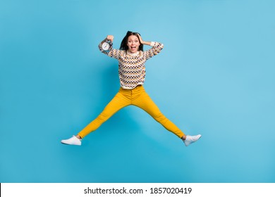 面白い若い女性の完全な長さの写真は、分離された時計の青い色の背景を保持している手の頭をジャンプ飾りセーターを着用します。