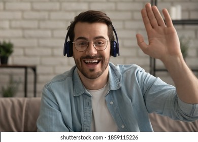 Visão da tela de tiro na cabeça de um jovem bonito, sorridente e sincero, de óculos e fones de ouvido, olhando para a câmera, fazendo um gesto de "olá, oi", dando boas-vindas aos amigos em uma reunião por webcam com vídeo online.