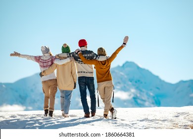 Cuatro amigos felices están de pie y abrazándose contra montañas cubiertas de nieve en un día soleado. Concepto de vacaciones de invierno