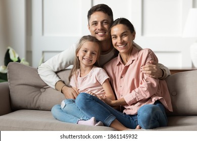 愛する若い男が愛する妻を抱きしめ、小さな娘がリビングルームのソファに座り、写真のポーズをとるカメラを見て微笑む幸せな人々。模範的な家族の肖像画、愛と絆の概念