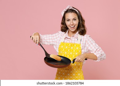 Smiling young woman housewife 20s in yellow apron checkered shirt making pancake with frying pan spatula while doing housework isolated on pastel pink background studio portrait. Housekeeping concept