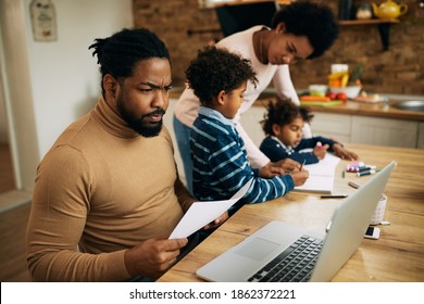 African American man using computer and working at home while his wife is homeschooling their children.