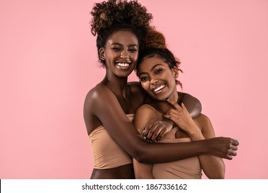 Excited emotional young women posing together , smiling and looking at camera. Pink pastel studio background. Beauty portrait of two black female models. Perfect toothy smiles.