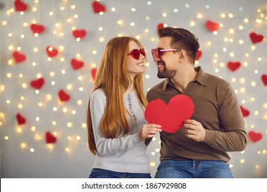 Happy young couple in love having romantic photo shoot on Saint Valentine's Day. Man and woman holding red heart shaped card, looking at each other and smiling sitting against blurred background