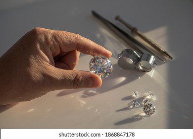 Close-up of the hand of jeweller gemologist with large size round cut diamond on desktop with magnifier and tweezers. Buyer diamond expert checking color of diamond in natural light.