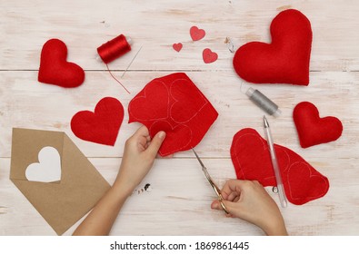 Vue de dessus d'une disposition de coeurs rouges cousus en feutre et de mains caucasiennes découpant des coeurs dans du feutre rouge avec des ciseaux sur un fond en bois blanc. Concept : cadeau fait main pour la Saint-Valentin.
