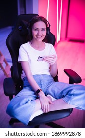 Gamer girl in a big professional music headphones in white t-shirt and blue jeans holds smart phone in hands and plays game app on mobile phone, smiling and looking at camera indoors.