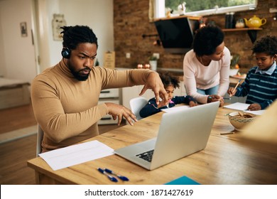 Black father using laptop and making video call while working at home. Mother is helping their kids homework in the background.
