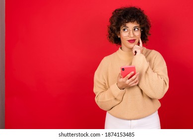 Denken dreemen Jonk schéin arabesch Fra, déi e gebreide Pullover droen, déi géint rouden Hannergrond steet mam Handy an d'Hand um Gesiicht hält. Entscheedungen huelen a soziale Medien Konzept.
