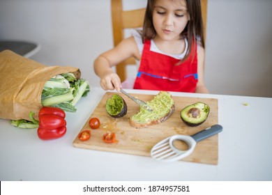 Cô bé đáng yêu trên bàn white trải bơ bên trên lát bánh mì. Trẻ nhỏ dễ mến và hạnh phúc cắt rau trên thớt. Con trẻ em chuẩn bị thức ăn