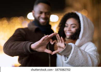 Te amo. Primer plano de una alegre pareja afroamericana positiva haciendo forma de corazón con los dedos caminando juntos por la noche, mirando las manos, enfoque selectivo, fondo borroso. Día de San Valentín
