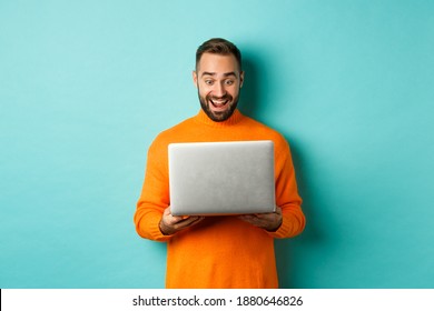 Hombre emocionado con suéter naranja trabajando en una laptop, mirando la pantalla de la computadora asombrado, de pie sobre un fondo azul claro