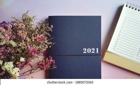 2021 blue note book or planner and calendar flat lay on pastel purple pink gradient background with blurred foreground of flower bunch. Hello new year, goal setting, start making resolutions theme.