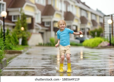 Little boy wearing yellow rubber boots jumping in puddle of water on rainy summer day in small town. Child having fun. Outdoors games for children in rain.