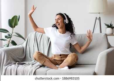 Bekymringsløs brunette kvinde lytter til musik og synger, bruger hovedtelefoner, sidder på sofaen i stuen, kopierer plads. Glad ung dame med lukkede øjne nyder musik, boliginteriør