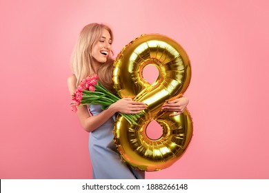Fröhliche junge Dame mit Blumenstrauß und goldenem Ballon Nummer acht zum Tag der Frau auf rosa Studiohintergrund. Reizende tausendjährige Blondine, die Frühlingsferien feiert