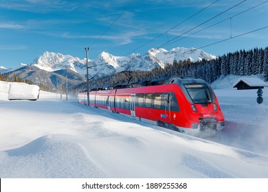 A local train traveling by a forest in a valley covered by deep snow on a sunny winter day and Karwendel Mountain dominating the background under blue sky near Garmisch-Partenkirchen, Bavaria, Germany