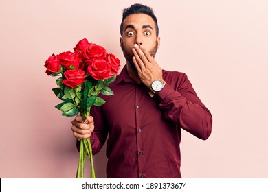 Joven hispano sosteniendo flores cubriendo la boca con la mano, conmocionado y asustado por error. expresión sorprendida