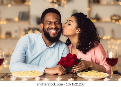 Mừng Ngày Đặc Biệt. Bạn gái da đen xinh đẹp hôn lên má bạn trai đang mỉm cười, tay cầm bó hoa hồng đỏ. Cặp đôi ngồi cùng bàn trong nhà hàng sang trọng trong buổi hẹn hò và ăn tối lãng mạn