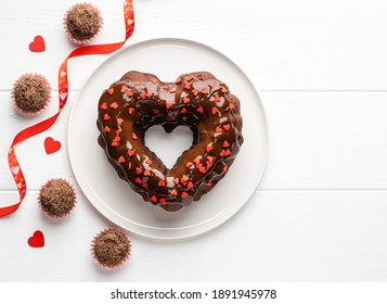 Schoko-Bundt-Kuchen in Herzform, bedeckt mit Schokoladenglasur und roten Streuseln und Schokoladentrüffeln. Dessert zum Valentinstag. Festliches Essen. Ansicht von oben. Platz kopieren. Weißer hölzerner Hintergrund.