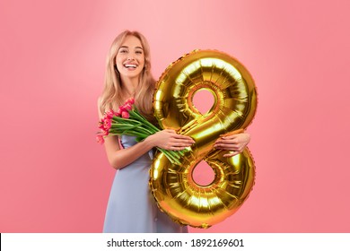 Charmante tausendjährige Dame mit Frühlingsblumen und goldenem Ballon, die den Tag der Frau auf rosafarbenem Studiohintergrund feiert. Herrliche junge Blondine, die Bündel Tulpen und Feiertagsdekoration hält