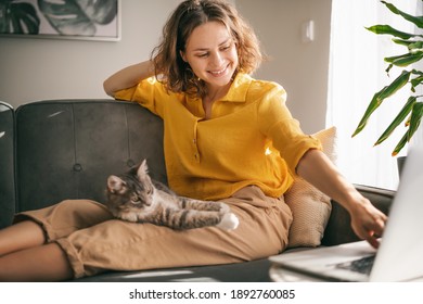Mulher casual de camisa amarela trabalhando no laptop com seu gato no sofá, sentadas juntas em uma sala com janela e planta verde.