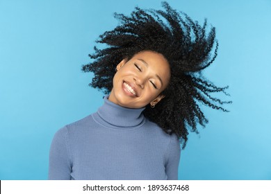 Feliz mujer afro sonriendo, tiene buen humor, bailando con su cabello rizado negro volando, aislada en el fondo azul del estudio. Chica afroamericana emocionada con peinado volador. Concepto de vida positivo.