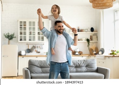 自宅でアクティブなゲームをプレイする父の首のピギーバックに座っている幸せな小さな娘。かわいい女の子を保持している笑顔のお父さんは、腕を伸ばして背中を運んでいます。週末を一緒に過ごす家族。