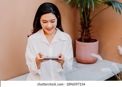 Asian woman playing games on smartphone while sitting on a concrete bench surrounded with pots of green plants with a desert sand colored wall in her background. Gaming addict outdoors