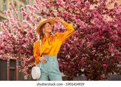 Glücklich lächelnde Frau mit trendigem Strohhut, gelber Satinbluse, blauer Hose, Cat-Eye-Sonnenbrille, mit Weidentasche, posiert auf der Straße in der Nähe von rosa Frühlingsblütenbäumen. Kopieren, leerer Platz für Text