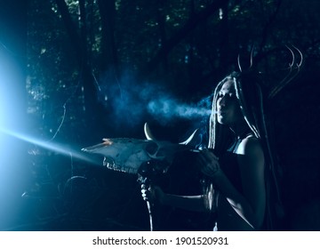 portrait of woman with long dreadlocks hair hold in hand staff with cow skull with horn against  wild forest trees Young girl smoke cigarette Woman shaman in ritual garment stand from fur and leather.