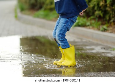 Little boy wearing yellow rubber boots jumping in puddle of water on rainy summer day in small town. Child having fun. Outdoors games for kids during rain.